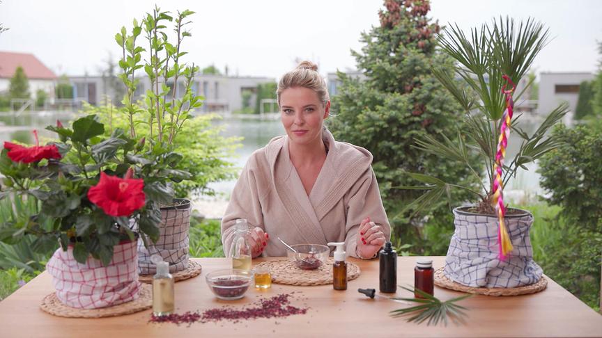 Christine Reiler im Bademantel demonstriert an einem Tisch mit einer Sägepalme und einem Hibiscus vor einem See die Zubereitung eines natürlichen Haarpflegeprodukts.