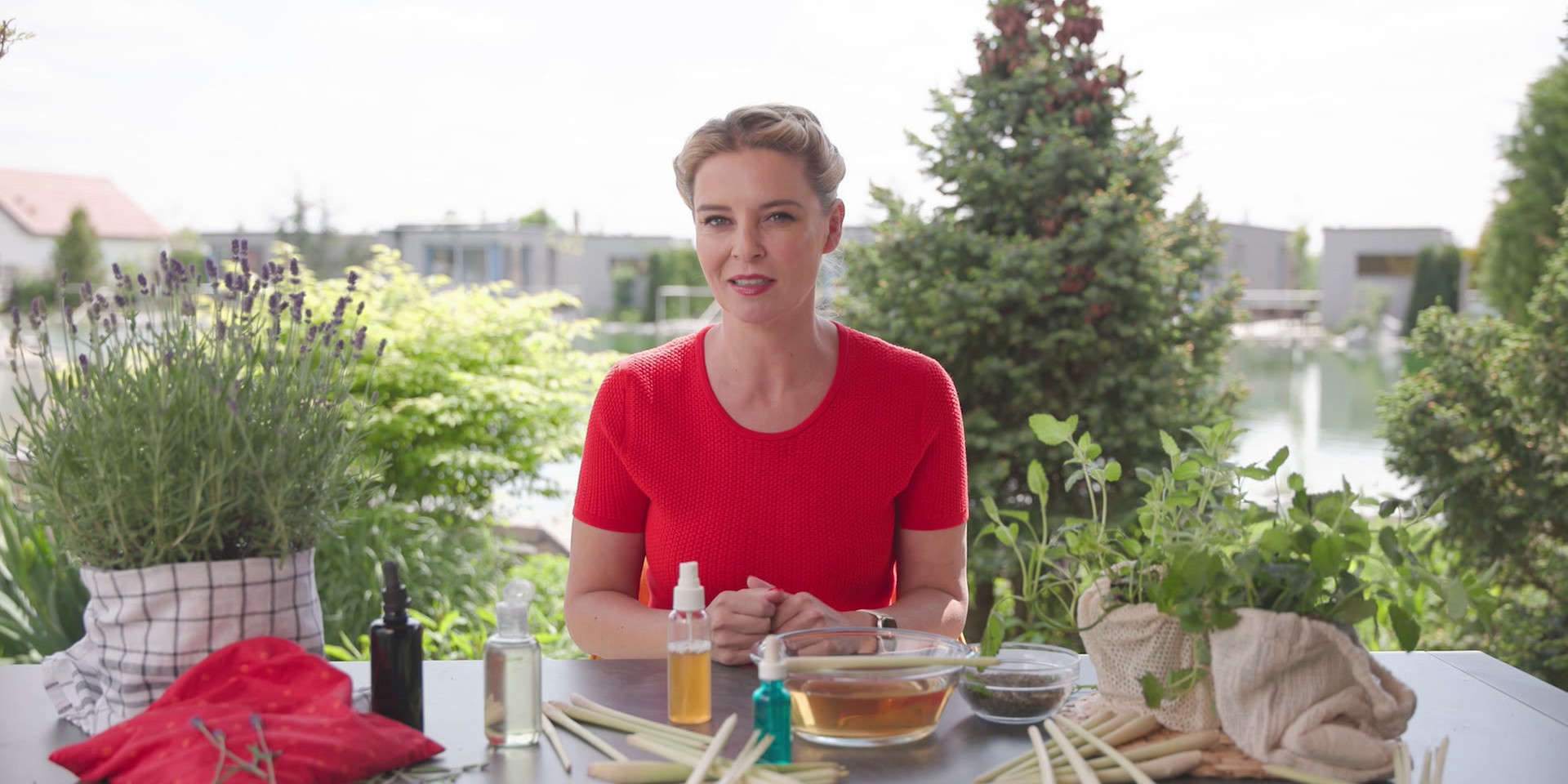 Christine Reiler sitzt outdoor an einem Tisch mit Kräutern wie Lavendel, Zitronenmelisse und Zitronengras, während im Hintergrund Gartenpflanzen zu sehen sind.