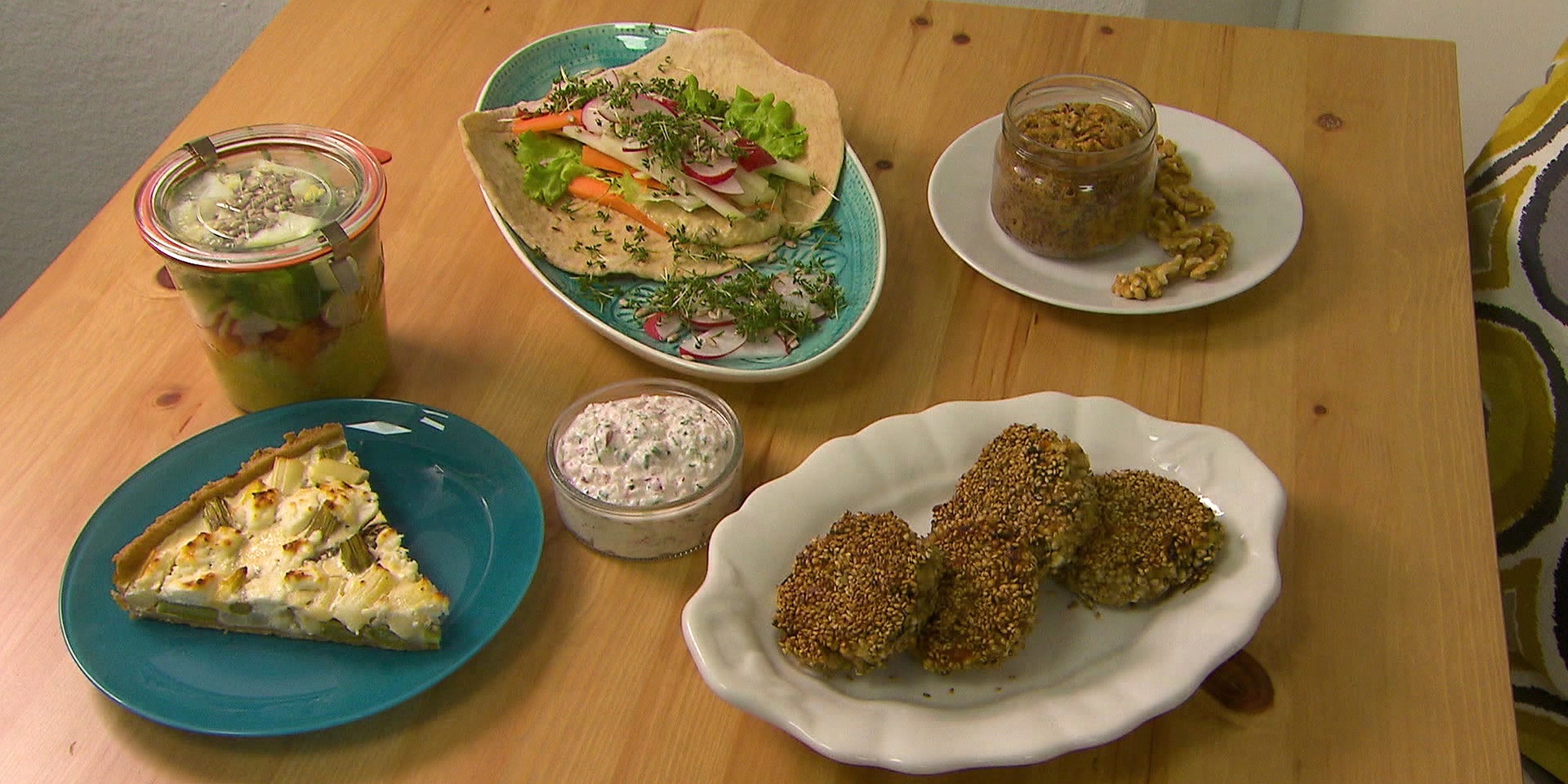 Auf einem Holztisch stehen eine bunte Mahlzeit: ein Glas mit eingelegtem Gemüse, ein Teller mit Wraps mit Hummus & Frühlingsgemüsefüllung, eine Quiche mit Spargel & Ziegenkäse auf einem blauen Teller, ein Karottenkuchen im Glas und Buchweizenlaibchen auf einem weißen Teller.