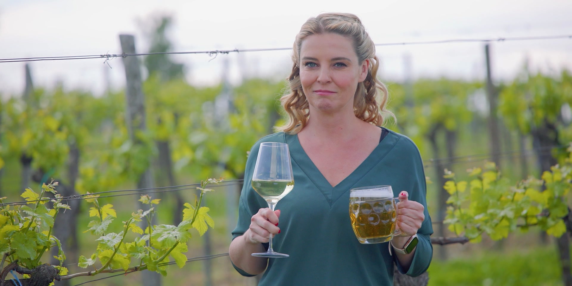 Christine Reiler hält in einem Weingarten ein Glas Wein und einen Krug Bier.