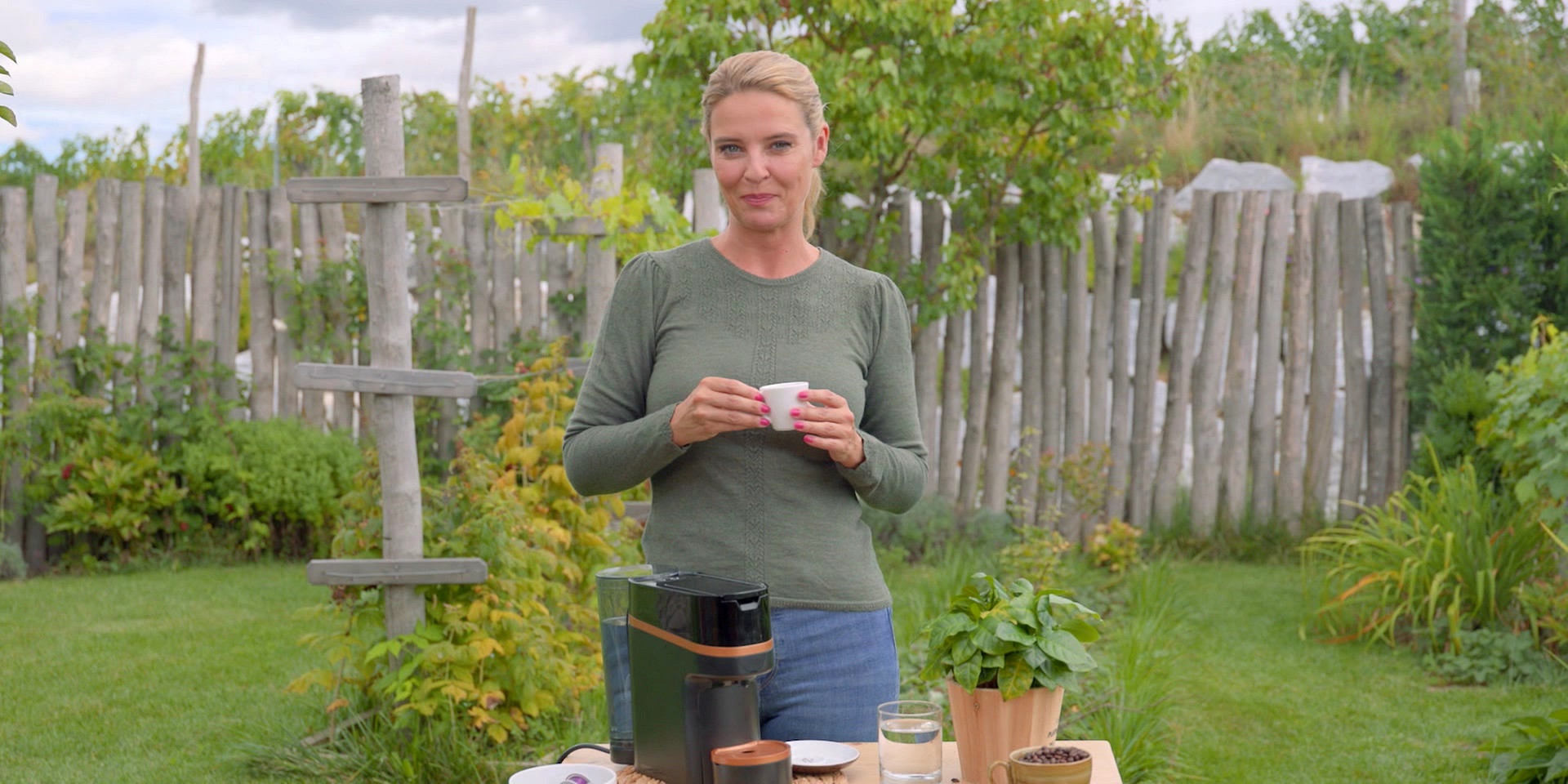 Christine Reiler steht im Garten neben einer Kaffeemaschine und hält eine kleine Espressotasse in den Händen.