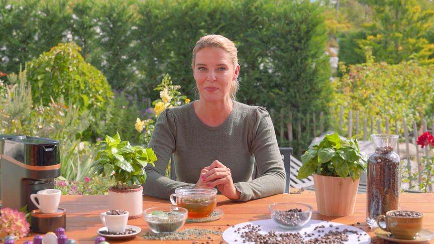 Christine Reiler sitzt in einem blühenden Garten an einem Tisch mit Kaffeemaschine, Kaffeebohnen und Pflanzen.