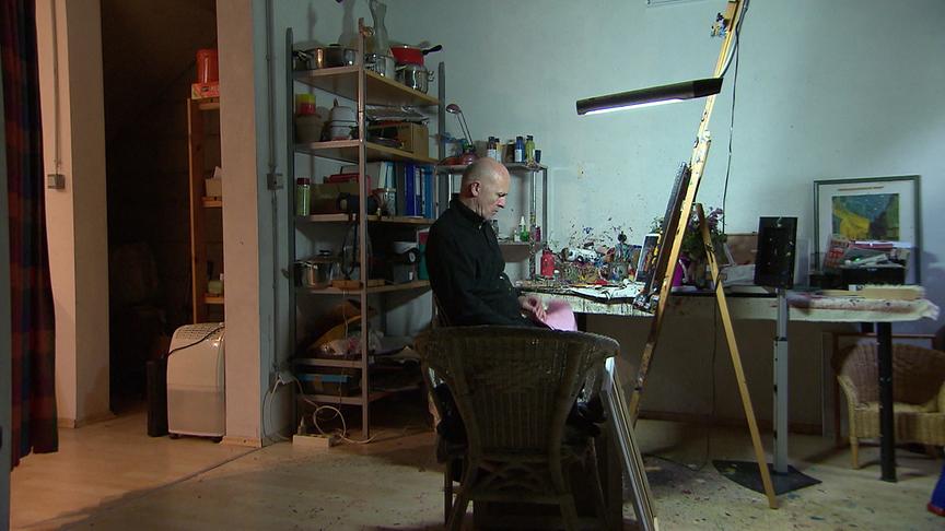 Tim Hind sitzt in einem vollgestellten Atelier an seinem Arbeitstisch vor einer Staffelei.