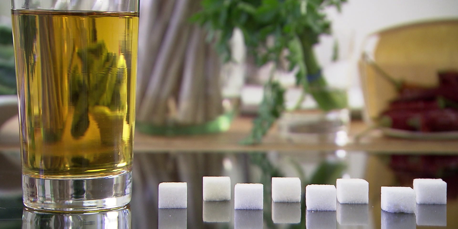 Neben einem Glas Tee liegen auf einer spiegelnden, glatten Oberfläche aufgereiht Würfelzuckerstücke.