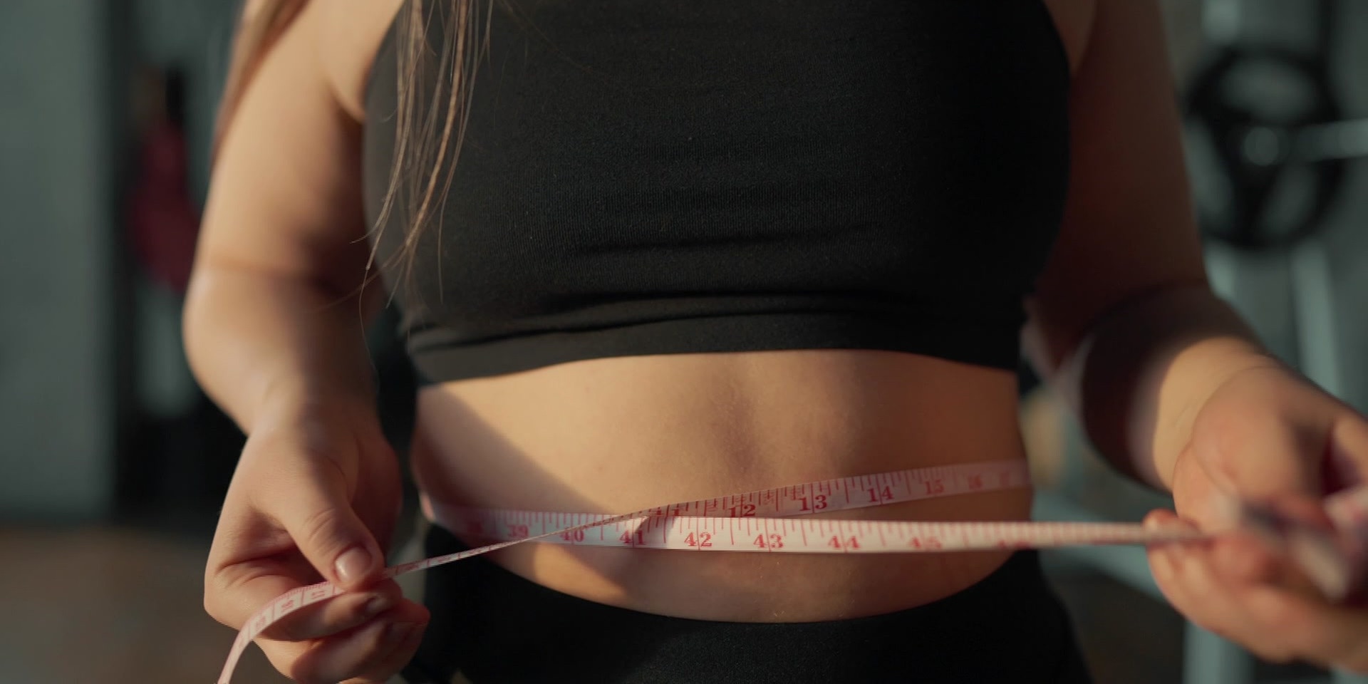 Eine Frau misst mit einem Maßband ihren Bauchumfang, während sie ein schwarzes Sporttop trägt.