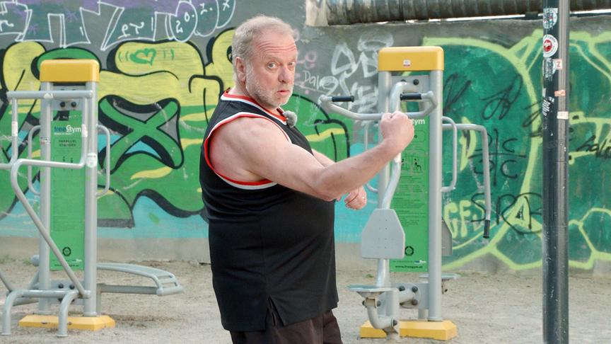 Reinhard Nowak trainiert an einem Outdoor-Fitnessgerät in einem Park mit Graffiti-Hintergrund und spannt dabei sichtbar seine Armmuskulatur an.