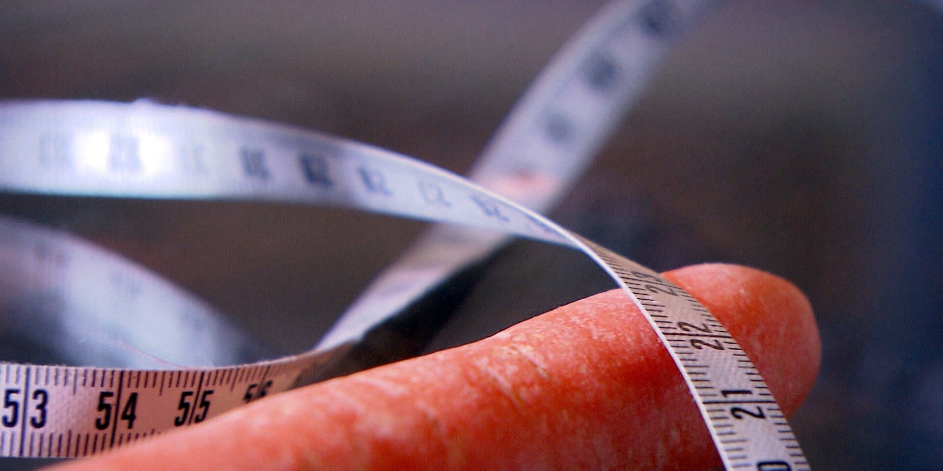 Ein Maßband liegt eng um eine Karotte gewickelt und symbolisiert das Abmessen von Körperumfang oder Größe.