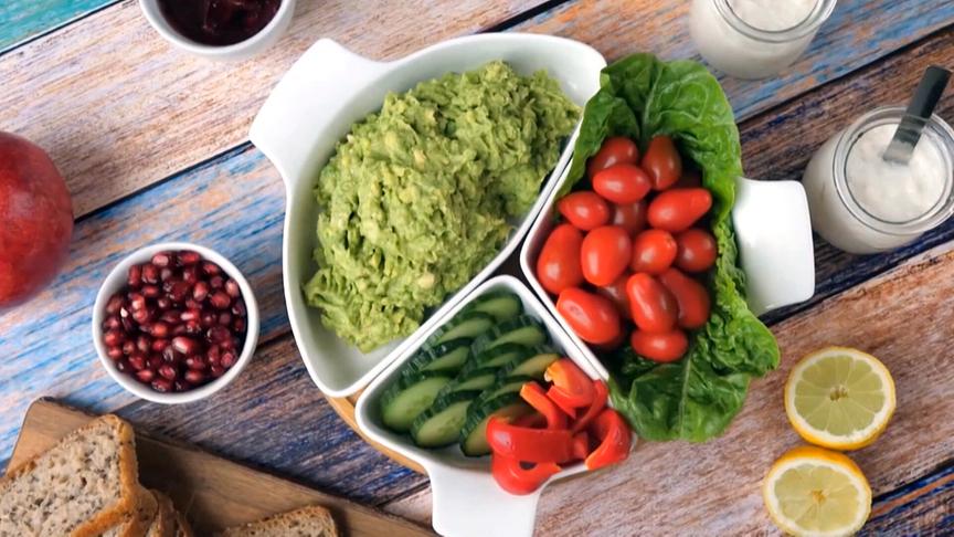 Eine farbenfrohe Auswahl frischer Lebensmittel wie Avocadocreme, Tomaten, Gurken, Paprika, Granatapfelkerne, Brot und Joghurt ist dekorativ in Schälchen auf einem Holztisch angerichtet.